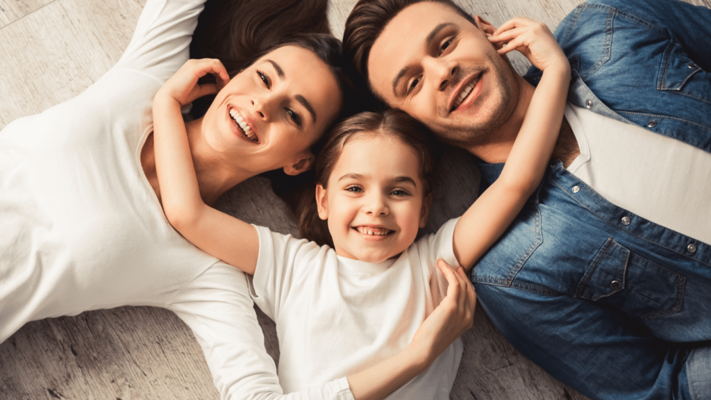 Happy family of three, with a little girl lying between her smiling parents, placing her hands on their cheeks.