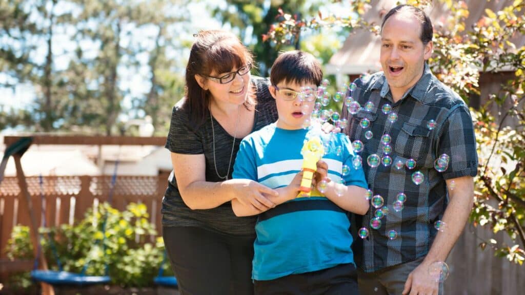 A young autistic boy shooting bubbles from a bubble gun, with his parents watching and smiling beside him.