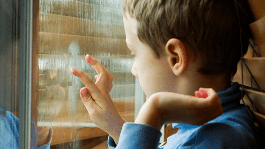 A young boy is drawing figures with his finger on a foggy window.
