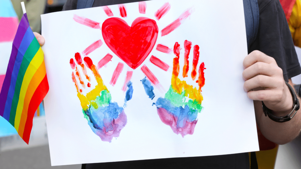 A person holds a piece of white paper with two rainbow handprints and a red heart painted on it. A rainbow pride flag and a trans pride flag are in the background.