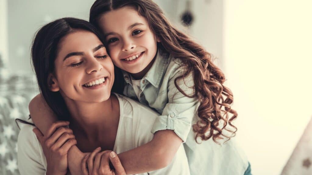 A daughter holding her mom from behind and both are smiling.