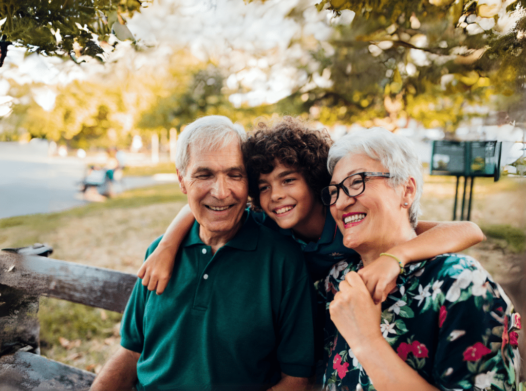 A smiling teenage boy with his arms around his two smiling grandparents. The grandparents are sitting on a bench in the park.