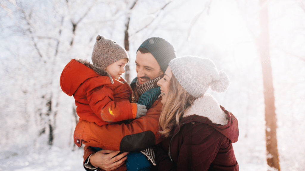 Family of three in winter outfits with snow in the backdrop. Dad holding the child in his arms while mom admiringly looks at the child, smiling.
