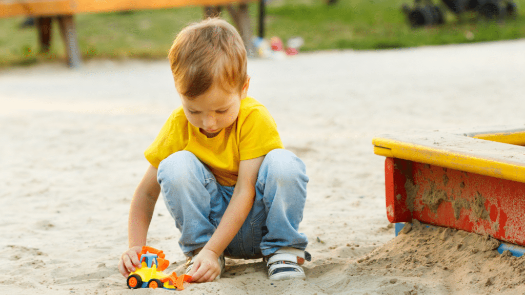 A blond kid, wearing a yellow T-shirt and jeans, is playing on the sand with a yellow and red truck.