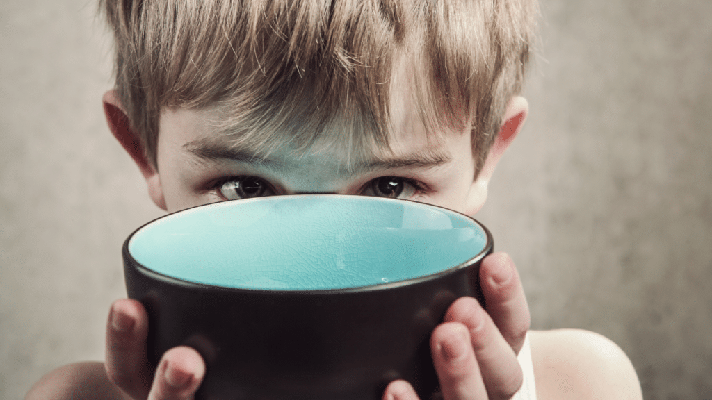 Blond boy holding a blue bowl in front of his face.