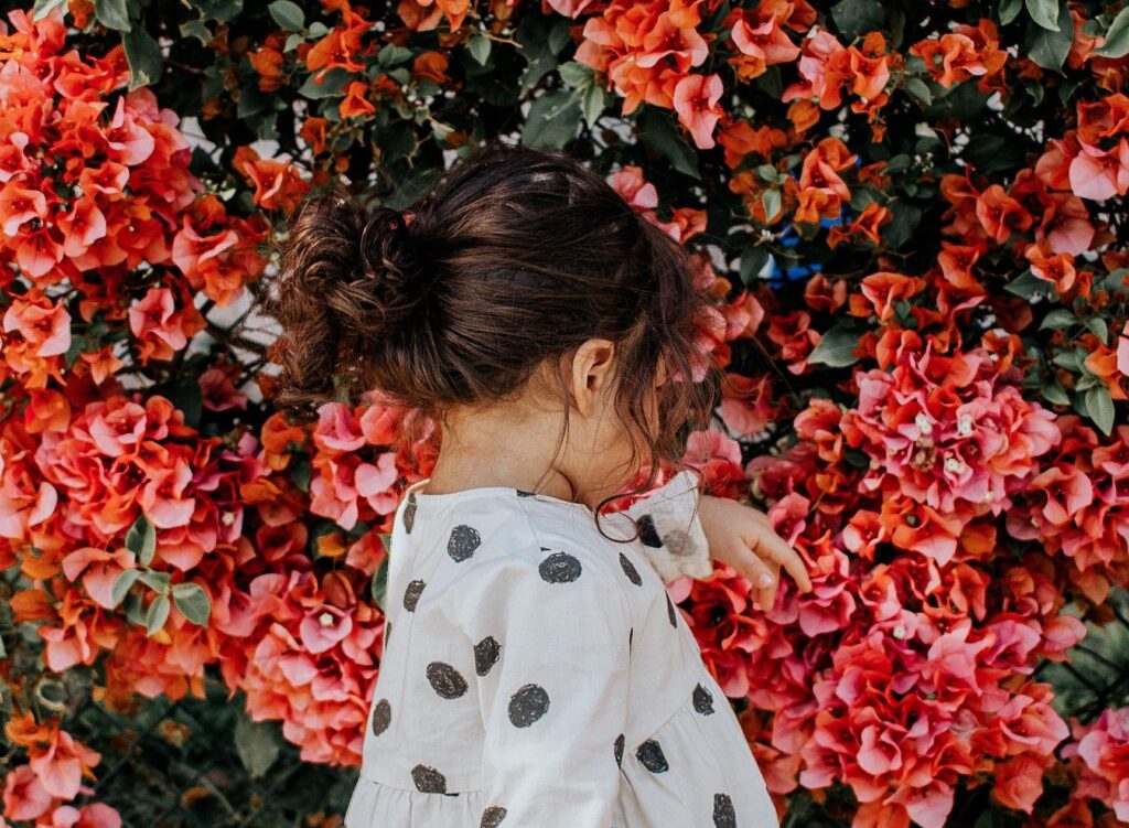 The back of a little girl wearing a white shirt with black dots. There are a lot of pink flowers in the background.