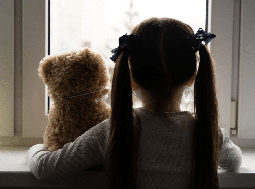 A little girl with two ponytails looking out the window and holding a teddy bear