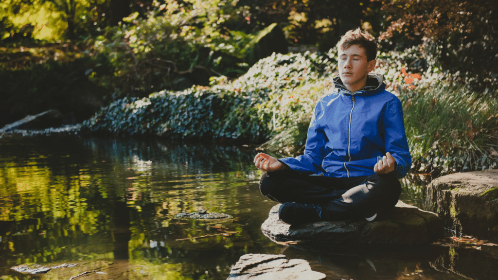 A male teenage boy meditating near a pond surrounded by greenery