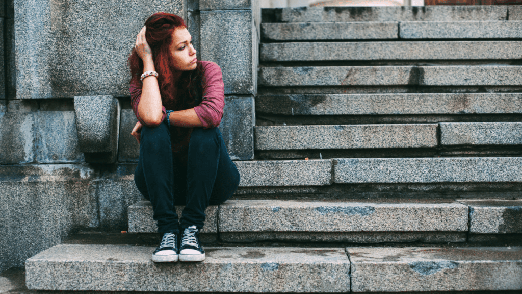 A teenage girl with pink hair and a sad expression is sitting on gray stone stairs.