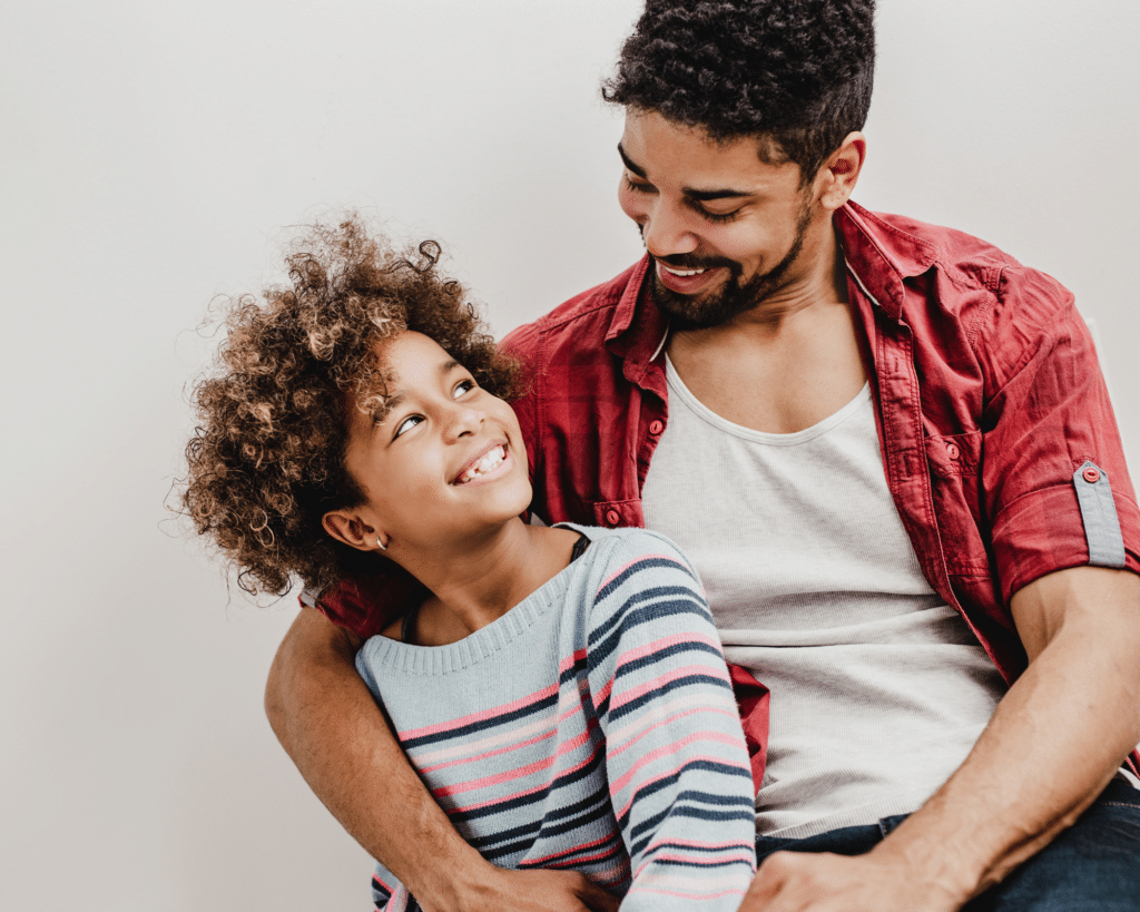 Happy African American girl with short curly hair and a bright smile looking at a man with brown skin and dark hair. The man is hugging and smiling at the child.