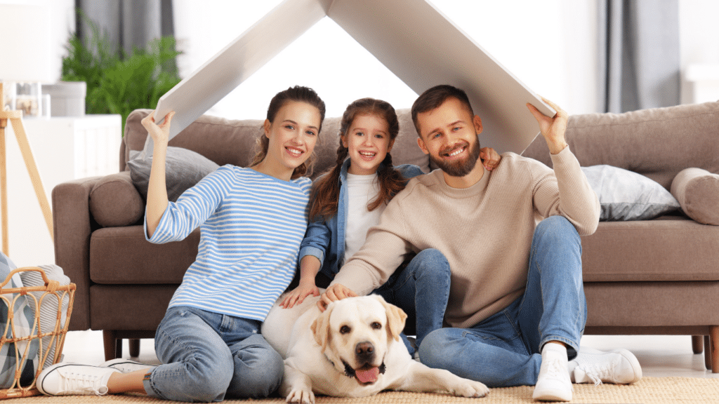 Happy family of three on the floor in front of a couch. Mom and dad hold a cardboard hut over their heads, creating a playhouse. Toddler daughter sits between them, and their dog is also present.