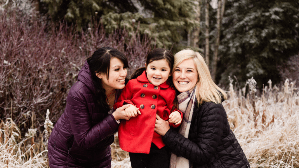 Picture showing the birth mother, the adoptive mother, and the child. The little girl, wearing a red coat, is positioned between both mothers, all of them smiling.