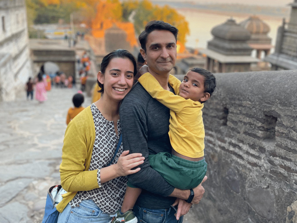 A mother and father, embrace each other and a little boy, their newly adopted son. They all have brown skin and black hair. They stand on a cobblestone street by a stone wall in India, where their son was born.