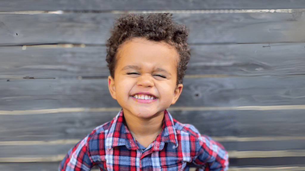 One of the Doornbos family members, a young boy with brown curled hair and dark skin, is smiling with his eyes closed due to the wide grin. He's wearing a shirt with red and blue stripes.