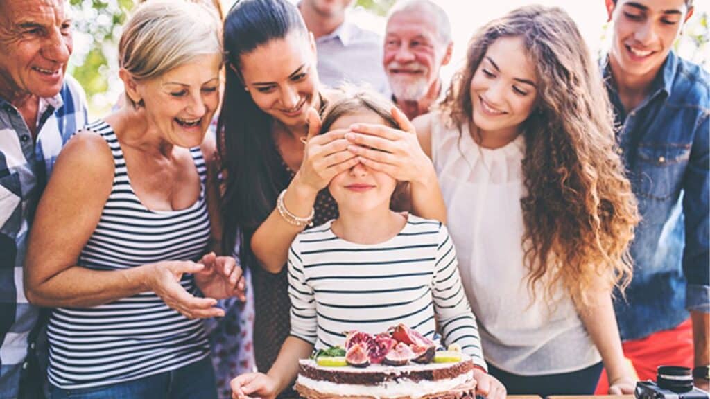 A young girl surrounded by a group of people, her eyes covered by a lady's hands, with a surprise birthday cake placed in front of her.