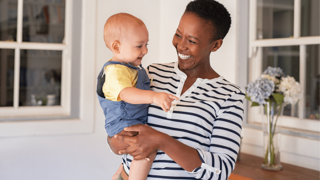 A black woman holding a baby in her arms and smiling while the child giggles.