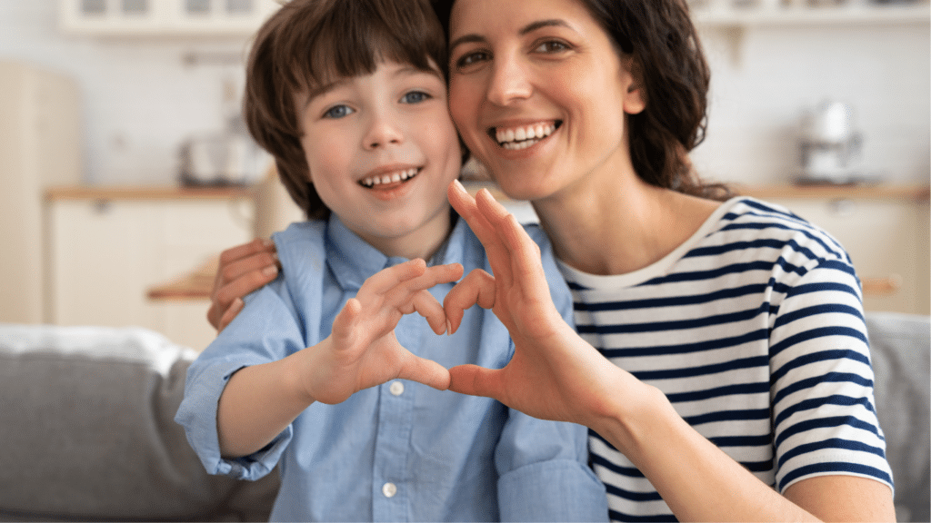 A mother and child, both smiling, attempt to create a heart shape with their fingers.