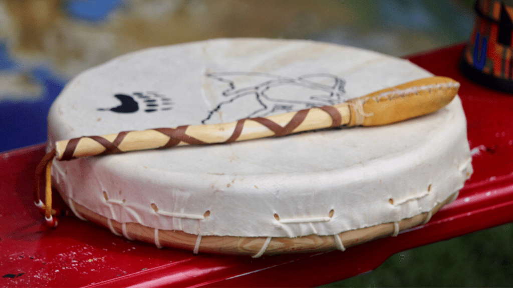 A small white Indigenous hand drum sits beneath a yellow drumstick adorned with red details.