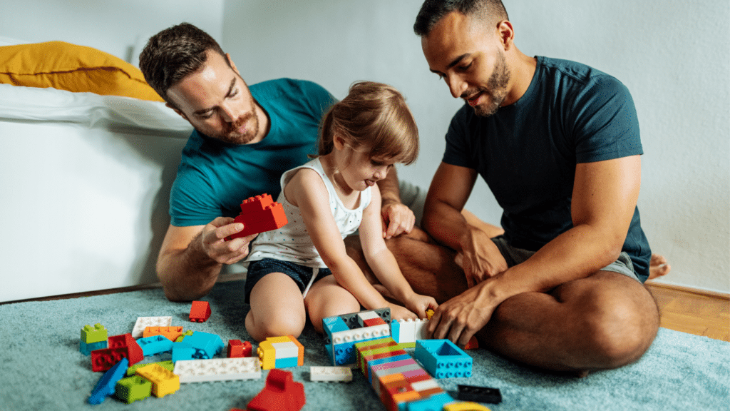 A gay couple fully engaged in playing with building blocks with a little girl.
