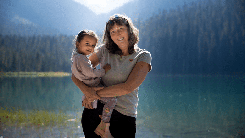 A woman holding a little girl in her arms, both smiling. In the background, there's a beautiful landscape of a lake and mountains.