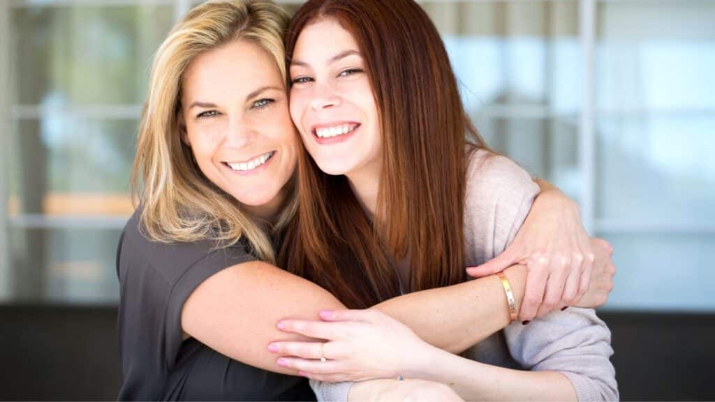 A teenage girl hugging her mother and both are smiling.