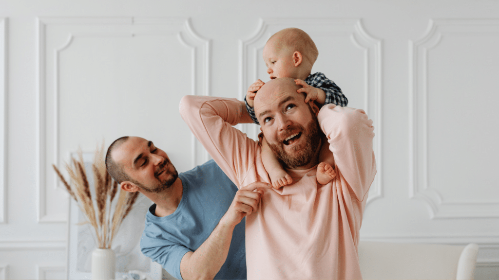 Gay couple playing with their child – one lifting the child above his head, the other playfully touching the child's toes.