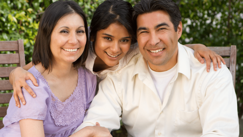 A teenage girl with her family, hugging both her parents and all three are smiling.