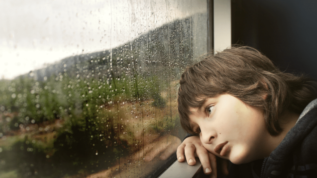A young boy with brown hair gazes out the window, appearing lonely and sad.