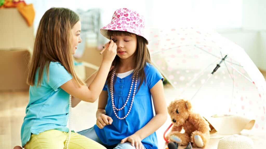 Two little girls sit on the floor with stuffed toys and an umbrella. They are dressing up with jewelry. One girl is putting eyeshadow on the other girl.