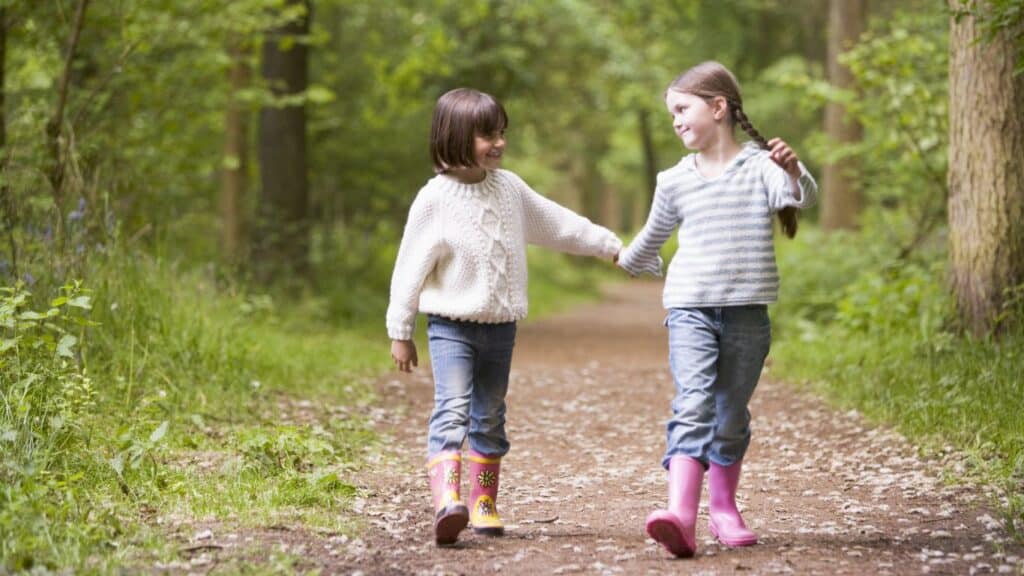 Two little girls in pink galoshes hold hands and smile at each other as they walk down a sunny woodland path.