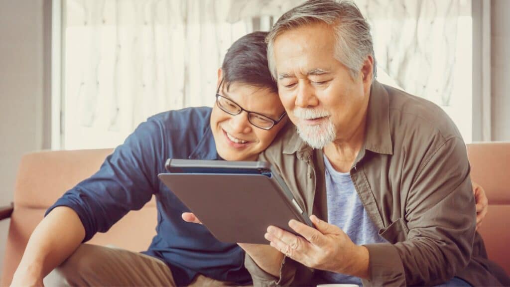 A young Asian man smiles and rests his head on his father's shoulder. They are sitting on a couch and looking at a tablet, as if learning about the process of adult adoption.