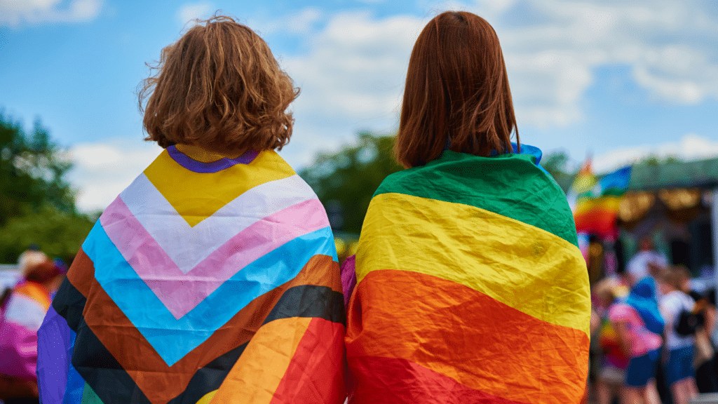 Two young adults with backs turned, draped in LGBTQ flags.