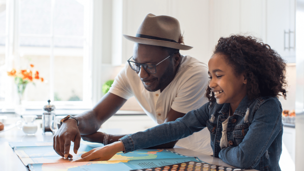 Black father and daughter sitting at a table, looking at a map, both smiling.