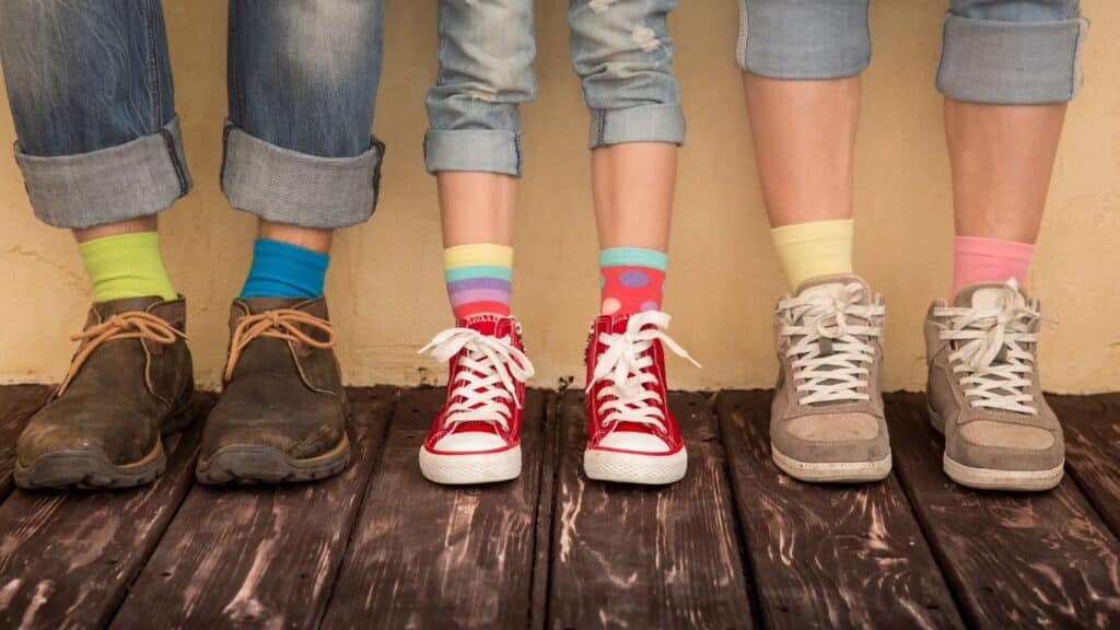 Three people with light skin are shown from the calf down. They are all wearing shoes, socks, and rolled-up jeans. The person in the middle is a teen wearing red shoes that are high-top sneakers. They also wear mismatched colourful socks. The adults on either side wear hiking shoes and their socks are also mismatched. Red shoes are a symbol of FASD with the slogan "Red shoes rock."