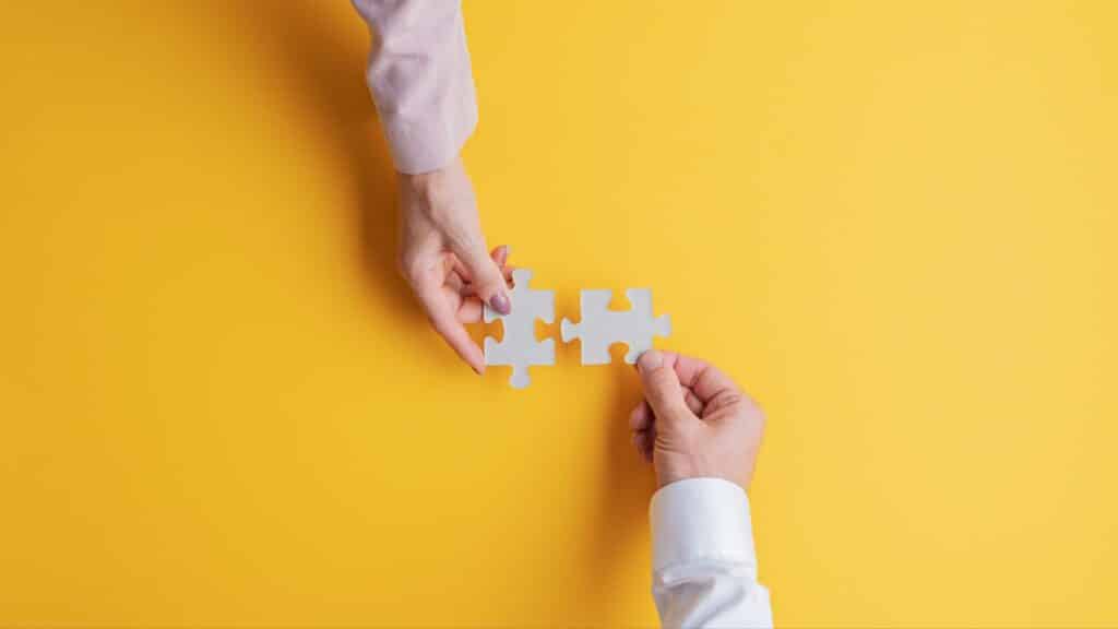 A woman's hand holding a white piece of a puzzle and a man's hand holding another piece, against a yellow background.