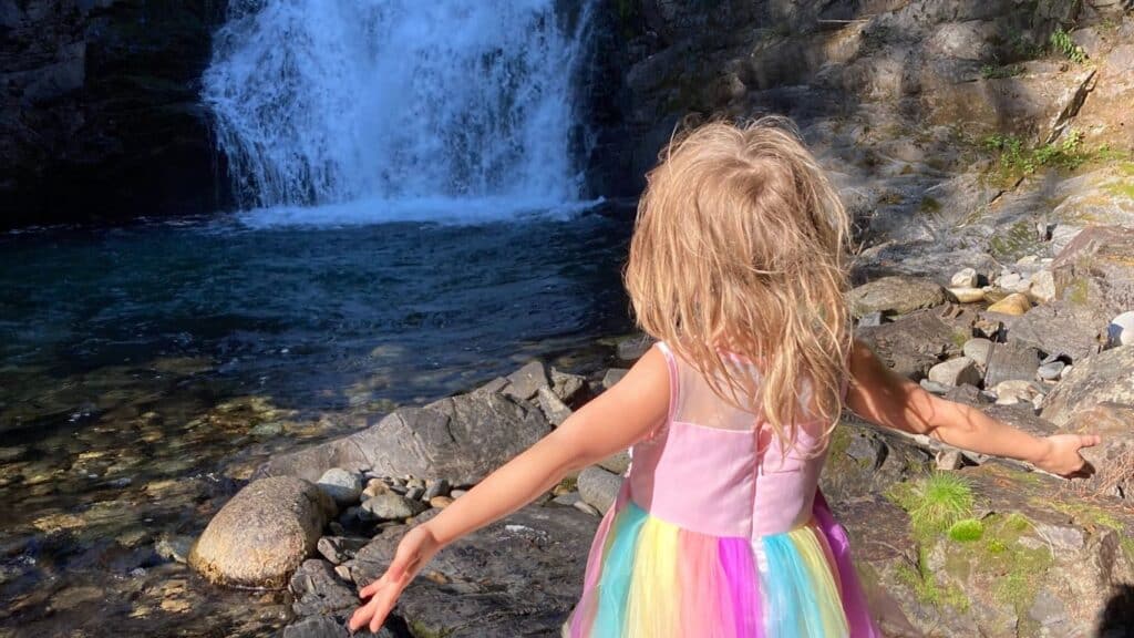 A little blond girl, seen from the back, wearing a rainbow-colored dress with her arms wide open. In front of her, there's a waterfall.