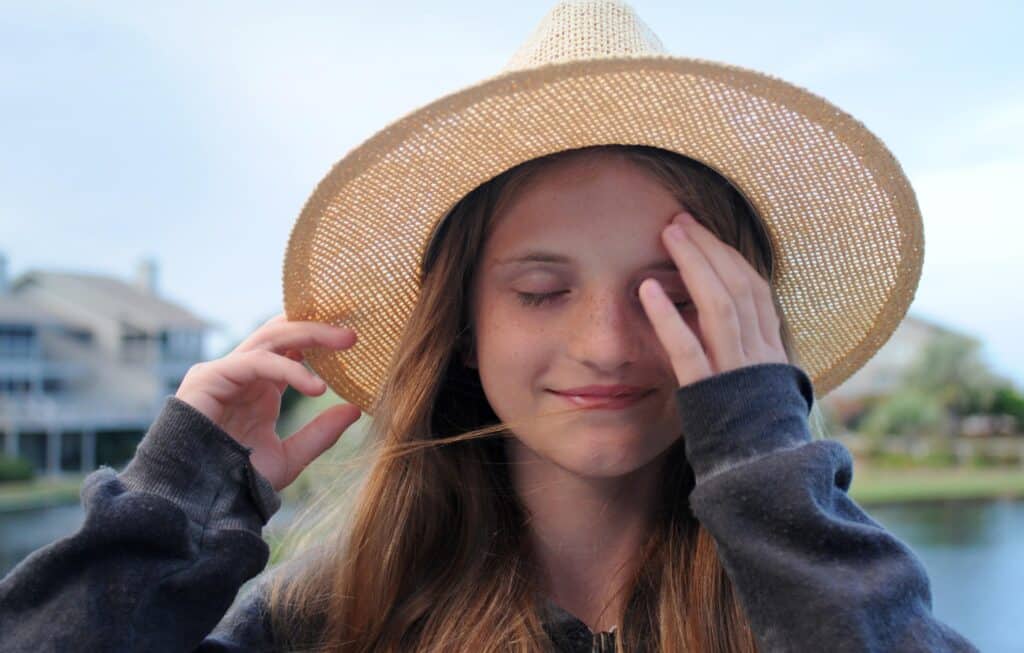 A young girl wearing a hat and smiling with her eyes closed, one hand covering half of her face.