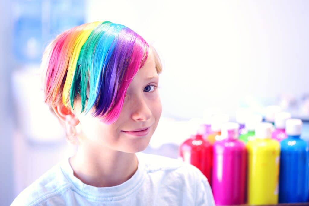 A child with rainbow-dyed hair smiles shyly at the camera.