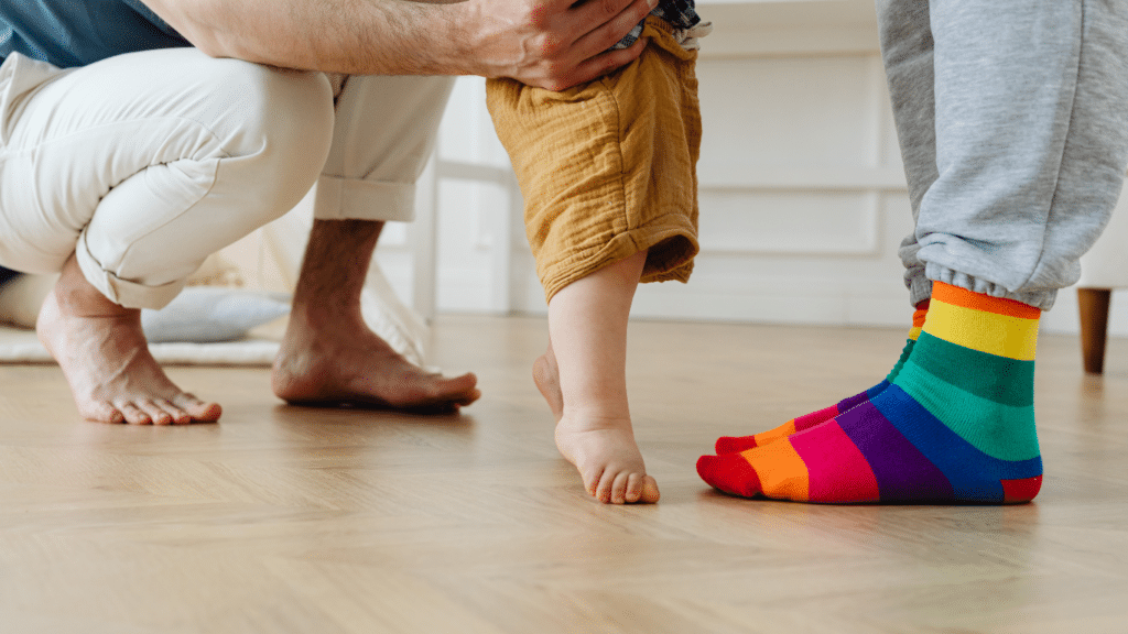 Feet of a child and two adults. One of the adults is wearing socks with the colors of the gay pride flag.
