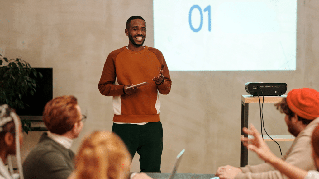 A young Black man stands in front of a small crowd. He is smiling and giving a presentation.