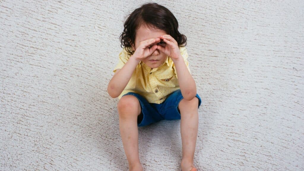 A young boy is sitting on the floor and pretending to use his hands as binoculars.