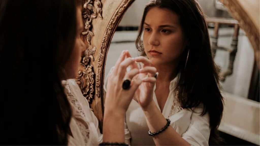 A young girl stands in front of a mirror, her reflection showing a sad and confused expression. She appears to be deep in thought, perhaps contemplating something troubling.