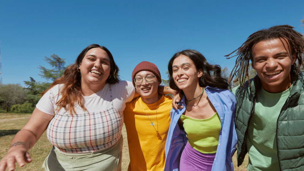 Four teens or young adults stand in a field with their arms around each other. They're all smiling. From left to right: a young East Asian woman with brown hair and skin; a young Asian man with glasses and a toque; a young woman with long brown hair and brown skin; a young Black man with dreadlocks.