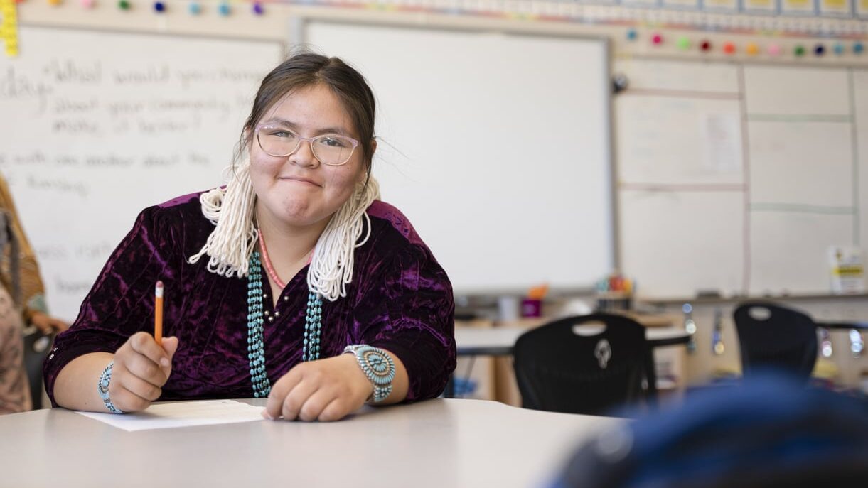A young Indigenous student sitting at a desk in a classroom, smiling at the camera