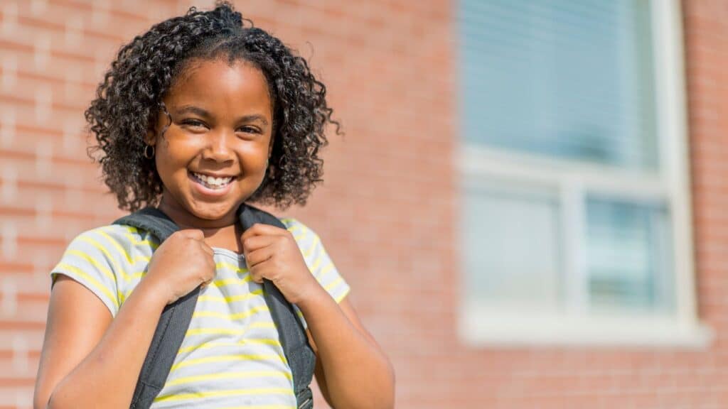 A girl smiling and holding onto the straps of her backpack