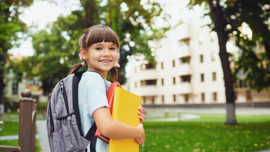 Girl smiling wearing a grey backpack and holding one yellow and one red binder