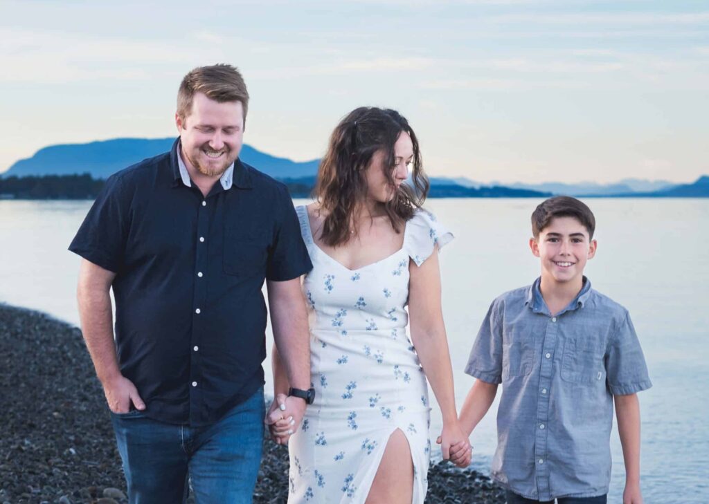 A father, a mother, and their young son hold hands and walk beside the ocean. They are all dressed in blue.