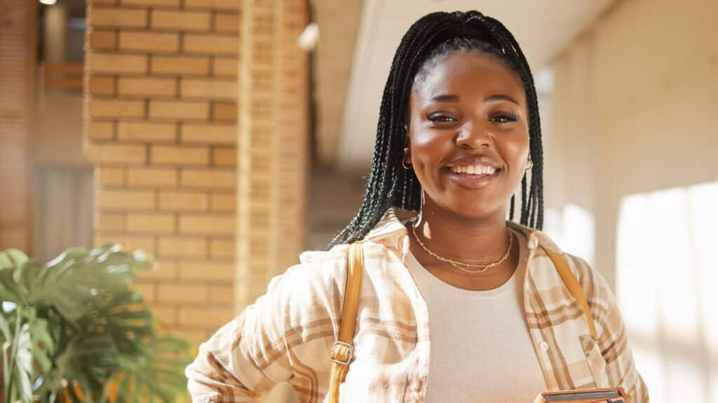 A smiling young woman standing in a hallway with a brick wall and green plant behind her.