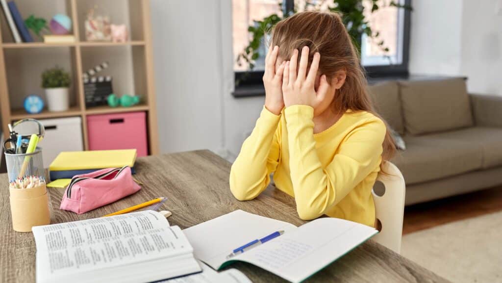 A young girl in a yellow shirt sits at a desk with her head in her hands. In front of her are open notebooks, a pencil, and school supplies.