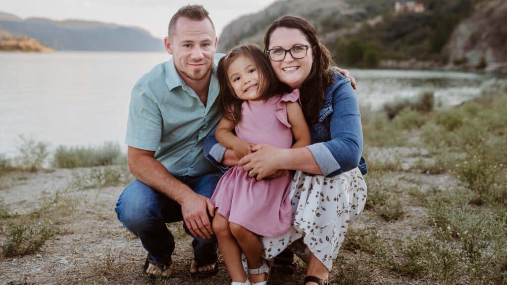 The Loeffler family smiling outdoors by a lake with mountains in the background.